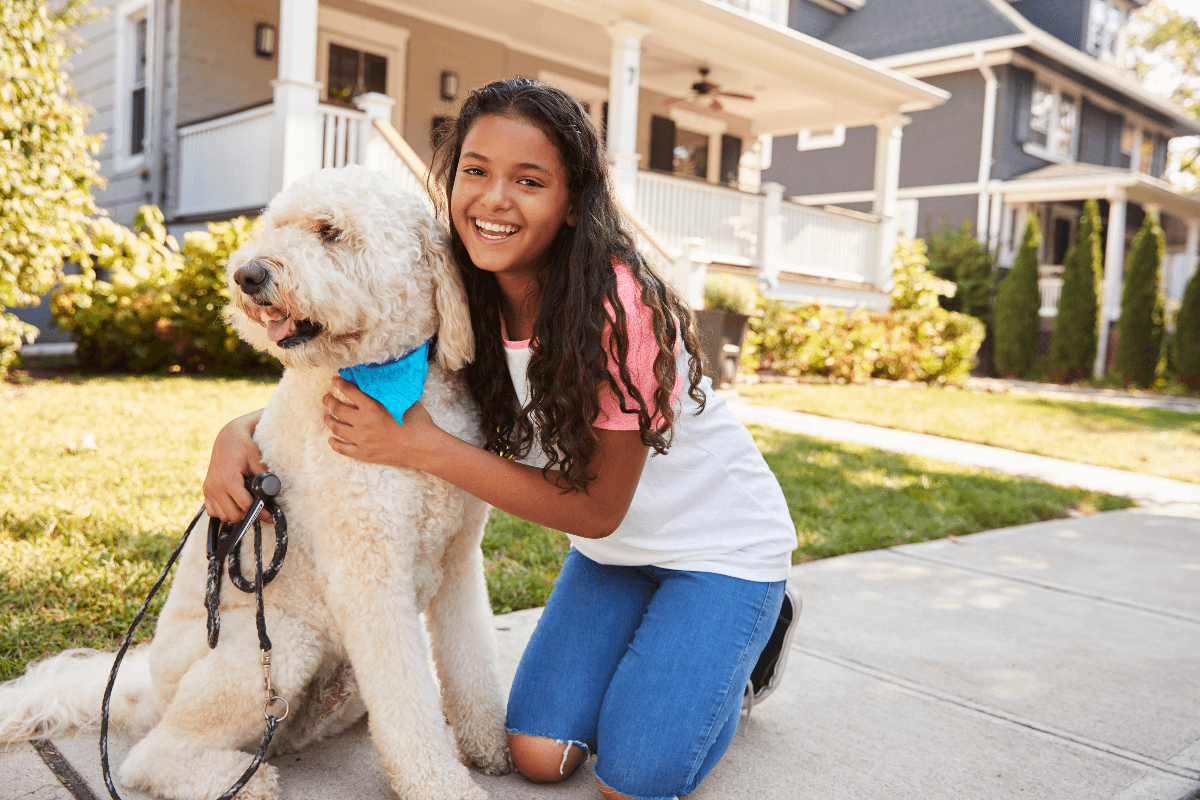 Young girl smiling and hugging her fluffy dog outside a suburban house.